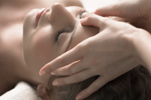 a woman lies on her back facing the ceiling on a massage chair. Her eyes are closed and she looks relaxed. A pair of hands massages between her eyebrows.