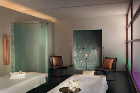 a massage room with two massage tables donned in white cloths. On the right table there is a platter with three glasses and a bowl. There is a soaking tub with two brown chairs on either side of the tub.