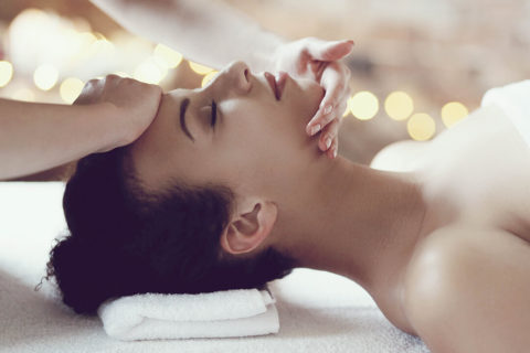 a woman with brown hair tied into a bun lays on her back on a massage table. Her head is propped up with a white towel. Her eyes are closed and she looks relaxed. A pair of hands massage her neck and forehead.