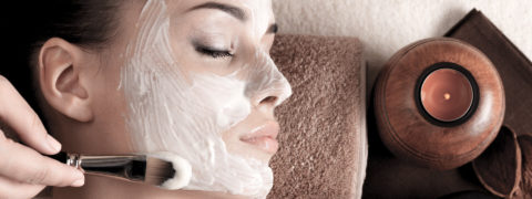 a woman with brown slicked back hair lies on her back on a massage table. Her head is propped up by a terrycloth towel and her head is tilted so her profile is showing. A pair of hands uses a brush to apply a white creamy face mask. There is a lit candle next to the woman's massage table.
