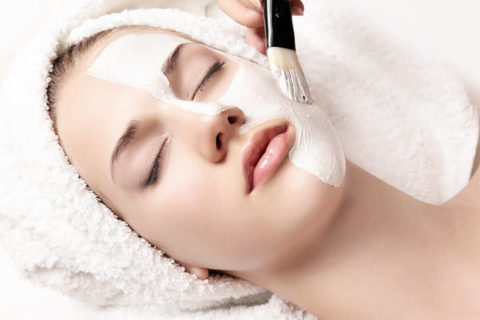 a woman with her hair wrapped in a towel lies on her back on a massage table. Half of her face is painted in a creamy white face mask. Her eyes are closed and she looks relaxed.