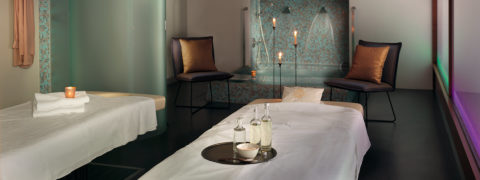 a massage room with two massage tables donned in white cloths. On the right table there is a platter with three glasses and a bowl. There is a soaking tub with two brown chairs on either side of the tub.