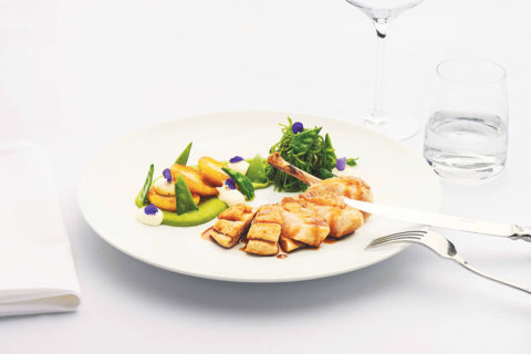 a detail shot of a beautifully prepared dinner. The plate is garnished with bright green herbs. Shiny silverware are leaned against the white plate. There is a glass of sparkling water next to the plate.