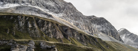 landscape shot of the Rhine Gorge. The mountain is layered with lush green grass on the base and gray, snow dusted rock at the peak. The sky is light gray with clouds.