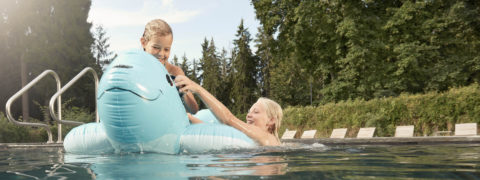 two young girls swim in the outdoor pool. One of the is sitting on a floaty shaped like a whale. The other girl is in the water trying to grab onto the floaty. In the background there are lounge chairs lining the pool. There are evergreen trees surrounding the area.