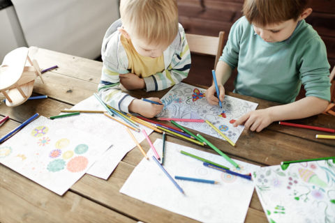 two little boys sit at a long wooden table. They are sharing a coloring page of flowers and butterflies. There are coloring book pages and colored pencils scattered around them.