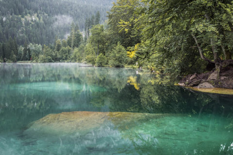 Landscape shot of Lake Cresta. The water is bright teal and clear enough to see the bottom of the lake. In the distance is an evergreen tree forest.