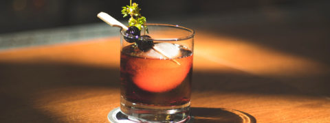 a detail shot of a craft cocktail. the glass is perched on a light wooden table. The cocktail is a deep red color. There is a single large ice cube floating in the glass adn the drink is garnished with blueberries and sprigs of green herbs