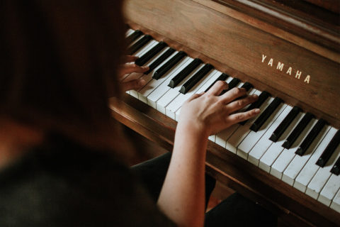 a detail shot of a woman hunched over a piano. Her fingers are extended on the keys. The piano is a dark wood Yamaha.