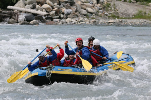a large blue and yellow raft boat carries a group of seven tourists and one guide through rapids in river. The water splashes up around them. They are all smiling and laughing.