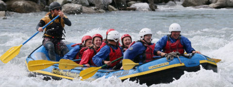 a large blue and yellow raft boat carries a group of seven tourists and one guide through rapids in river. The water splashes up around them. They are all smiling and laughing.