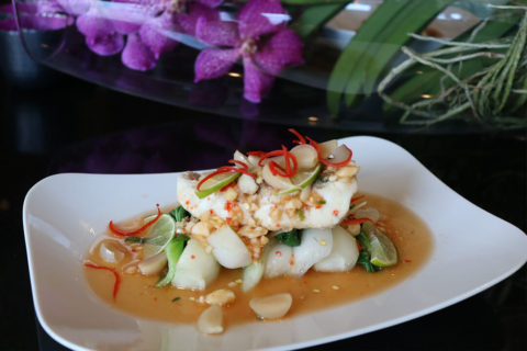 A detail shot of Thai dinner dish. The dish is artfully placed on a white plate. There are red and green garnishes atop the food. In the background are purple flowers.