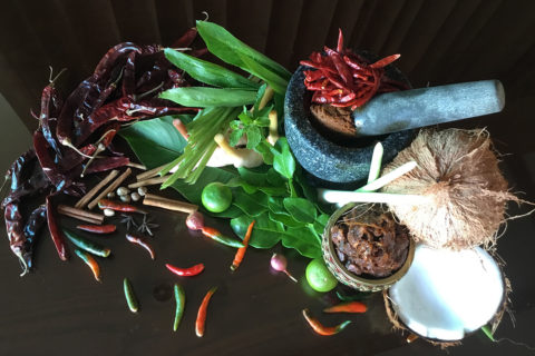 detail shot of thai spices placed on a dark brown table. The spices are bright greens and reds. There are peppers being ground in a pestle and mortar and there is a coconut split in two.
