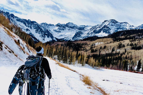 a man in snow gear walks on a snowy path. He is wearing a large gray backpack with two sets of snow shoes dangling from it. He is walking towards a forests of light and dark green trees. In the distance are snowy mountain peaks.