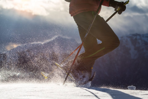 a close up shot of a snow shoer's legs are the step through the snow. Snow particles spray up around his feet as he moves forward. He holds poles in each hand and wears dark green snow pants and a red puffy jacket.