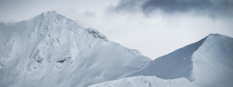 landscape shot of mountain peaks. The mountains are covered in a thick layer of powdery snow. There is a single dark cloud weaving through the two peaks. The sky is a light gray