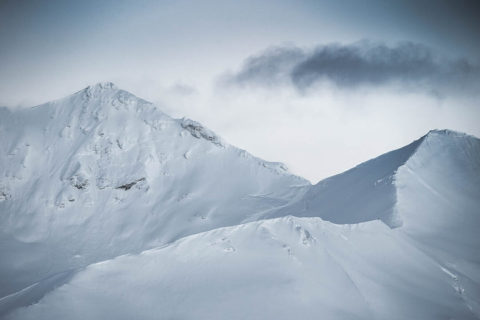 landscape shot of mountain peaks. The mountains are covered in a thick layer of powdery snow. There is a single dark cloud weaving through the two peaks. The sky is a light gray