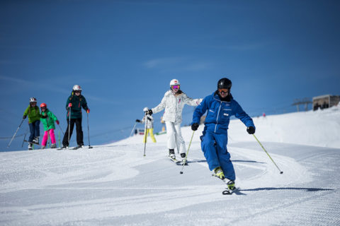 a ski instructor in an all royal blue outfit leads a line of children down a well groomed slope. The sky in the background is a deep, clear blue.