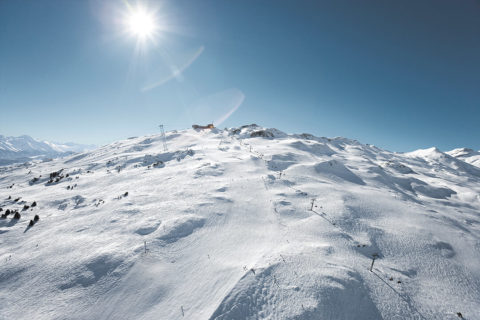 an aerial shot of a swiss ski resort. There is a long ski life running the length of the mountain. Little forms of skiers dot the groomed trails. Evergreen trees dot the mountain. The ski is clear and bright blue.
