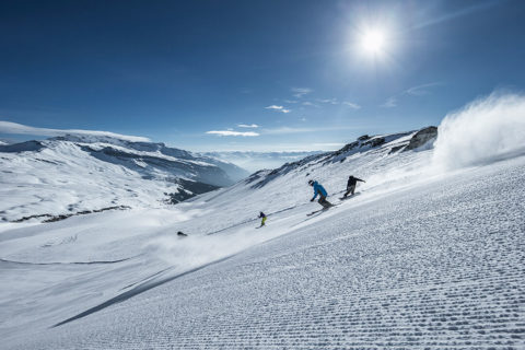 a group of skiers in bright, colorful outfits ski and snowboard down a slope. The slope is well groomed. In the distance are the jagged snow covered tops of the swiss alps. The sky is a bright blue with a few little clouds.