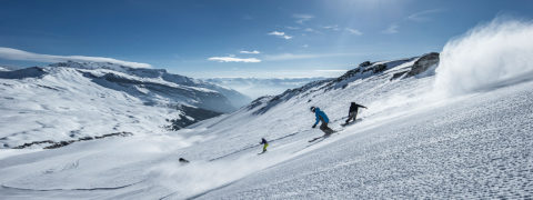 a group of skiers in bright, colorful outfits ski and snowboard down a slope. The slope is well groomed. In the distance are the jagged snow covered tops of the swiss alps. The sky is a bright blue with a few little clouds.