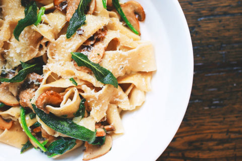 a detail shot of a pasta dish. The thick strands of pasta intertwine with golden mushrooms and bright green garnish. The pasta is sprinkled with little flakes of Parmesan cheese. The white bowl sits atop of a dark wood table.