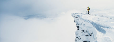a skier in a bright yellow jacket looks out over a cliff. The background of the photo is covered by white clouds.