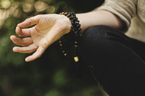 a detail shot of a woman's hand as it rests on her thighs while she sits cross legged performing yoga. Her palms are facing upwards. She has a dark beaded necklace wrapped around her wrist as a bracelet.