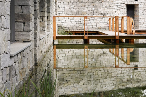 detail shot of the pond outside of the waldhaus flims spa. The water is glassy and reflects the stones of the walls surrounding it. There is a wooden platform dock that allows people to stand and observe the pond.