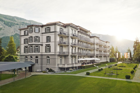 exterior shot of the Waldhaus Flims resort building and the surrounding grounds. The building is white with gray shutters. There are balconies at each window. The grounds are lush green grass with manicured bushes. In the background are the swiss alps.