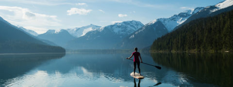 a woman in a pink workout shirt and black leggings stands on a paddle board. She uses a paddle to push her through the turquoise waters of Lake Cauma. In the distance are evergreen trees and the snow capped tops of the swiss alps.