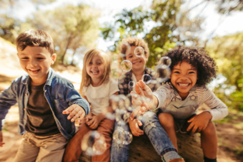 a group of four toddlers sit on a log. They are laughing and reaching toward a cluster of bubbles that are in the foreground