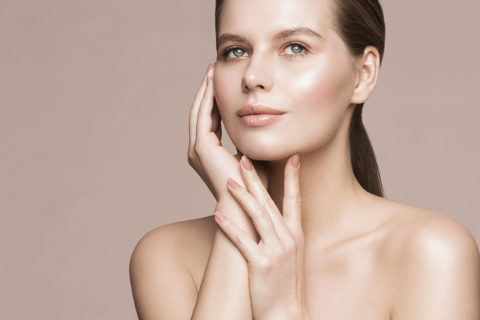 a woman with brown hair tied into a low, sleek ponytail caresses her face with her manicured hands. She is looking up over the camera and has a relaxed expression on her face. She is wearing a soft pink nail color and natural, glowy makeup