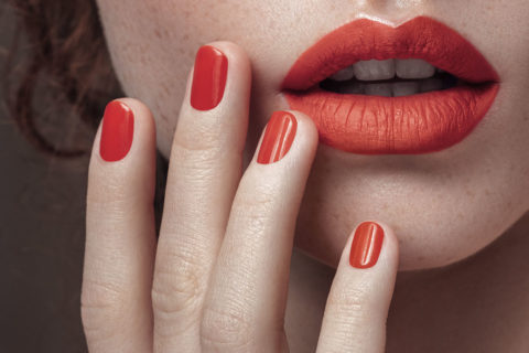 a close up of a woman's mouth. Her lips are slightly parted and are painted with red lipstick. Her hand is reaching up to touch her jawline. Her manicured hands are painted with red nail polish to match the color of her lips
