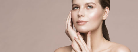 a woman with brown hair tied into a low, sleek ponytail caresses her face with her manicured hands. She is looking up over the camera and has a relaxed expression on her face. She is wearing a soft pink nail color and natural, glowy makeup