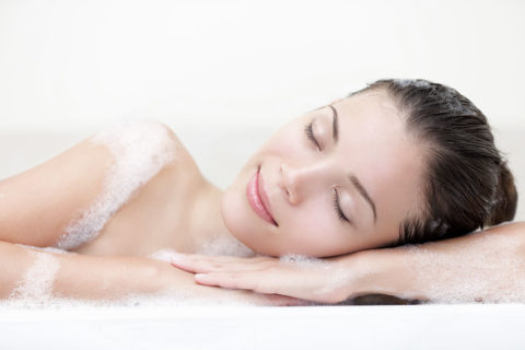 a woman lies on her side resting her head on her crossed forearms. Her eyes are closed, her damp hair is pulled back and she looks relaxed. She is covered in white bubbles from the bath she is soaking in.