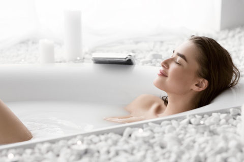 a woman with brown hair soaks in a bath. Her head is resting on the edge of the tub, her eyes are closed and she looks relaxed. She is surrounded by white rocks and white candles.