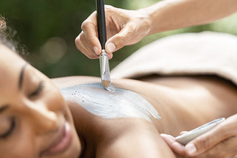 a woman lays face down on a massage table. He head is tilted towards the camera and her eyes are closed and she looks relaxed. A hand overhead uses a brush to apply a gray clay paste to the woman's bare back