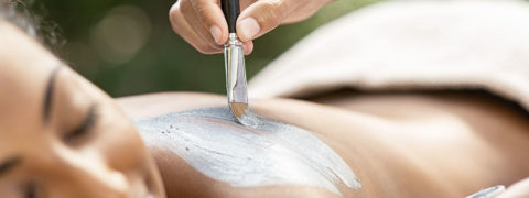 a woman lays face down on a massage table. He head is tilted towards the camera and her eyes are closed and she looks relaxed. A hand overhead uses a brush to apply a gray clay paste to the woman's bare back