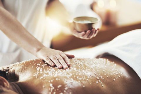 a woman lies on her back on a massage table. A massage therapist holds a gold bowl of salt crystals. She uses her hand to spread the salt crystals onto the woman's bare back.