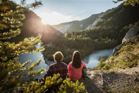 2 kids sitting on rock looking out to Waldhaus Park's mountain and lake, clear blue sky and sun glare
