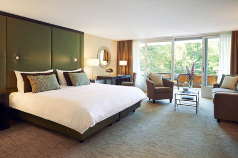 bedroom with full wall of window looking out at trees, a glass sliding door to the balcony, white bed with sofa, chair and coffee table