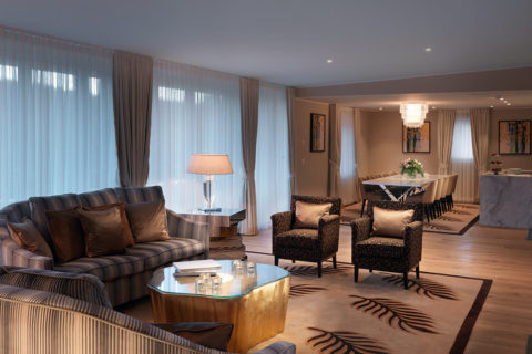suite living room with view of the Swiss mountains, comfy sofas, lamps, large windows, glass french door opens to balcony with lounge chairs, table with champagne