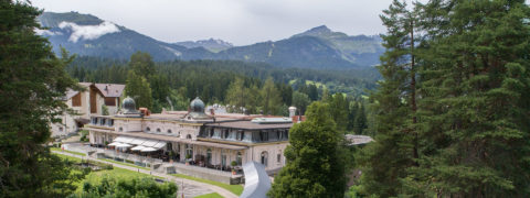 a landscape shot of the Waldhaus Flims resort building and the surrounding grounds. The building is white and is contrasted by the bright green of the evergreen trees that surround it.