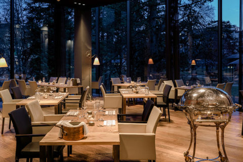 an interior shot of a formal dining room. There are multiple rectangular wooden tables around the room surrounded by white or black leather arm chairs. There is a grand silver serving cart in the corner of the room. The floor to ceiling windows allow for a view of the resort grounds at dusk.