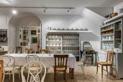 An interior shot of a white dining room. There is decorative silverware lining the shelved on the wall. There are three long tables donned in white linens. Lining the tables are white and brown alternating chairs.