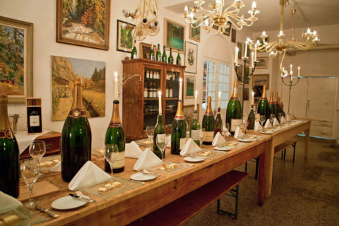 an interior shot of a dining room. There is a long wood table in the center of the room with long wood benched lining it. On the table are glass plates with slices of bread an cheese on them. There are different sized green glass bottles running through the middle of the table. On the wall are landscape paintings and a wooden cabinet full of wine bottles.