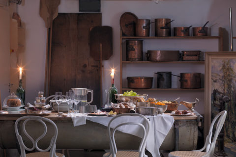 a private dinner table set up in the wine cellar. On the wall are copper pots lined on the wooden shelves as decoration. The long table is full with bowls of bread, grapes and cheeses. There is a bottle of wine in the middle of the table next to a tall candlestick. Plates and wine glasses are stacked on one end of the table across from the food. White chairs line the table.