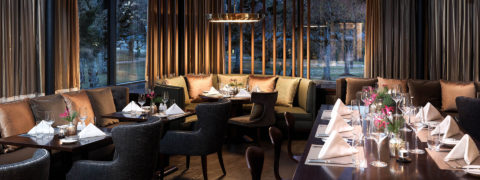an interior shot of the Siam dining room. There are floor to ceiling windows that allow for a view of the resort grounds. Allow the edges of the dining room are long couches with neutral colored pillows. Black tables line the couches and on the other side of the tables are charcoal gray chairs. In the center of the room is a long wooden table with pink and green flowers as the centerpiece.