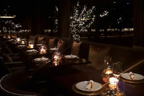 an interior shot of the Siam dining room. There are floor to ceiling windows that allow for a view of the resort grounds at night with the trees lit with twinkle lights. Allow the edges of the dining room are long couches with neutral colored pillows. Black tables line the couches and on the other side of the tables are charcoal gray chairs. There are candles in the center of every table.